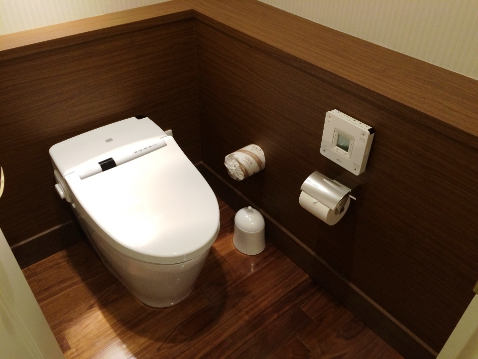 木目調の壁と床に囲まれた、清潔な白いトイレ。トイレットペーパーホルダーと壁の操作パネルが見える。